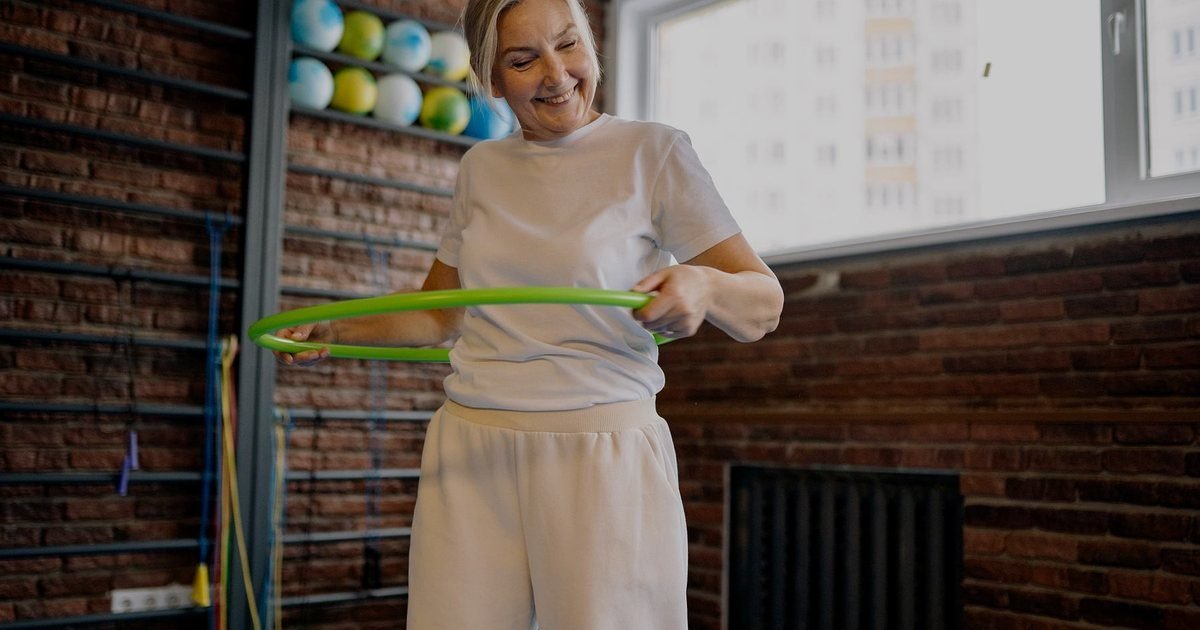 Mulher sorrindo segurando um elástico verde para treino em uma sala com tatame azul