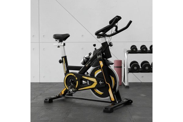 Bike de spinning WCT Fitness preta e amarela em uma sala de treino com halteres ao fundo