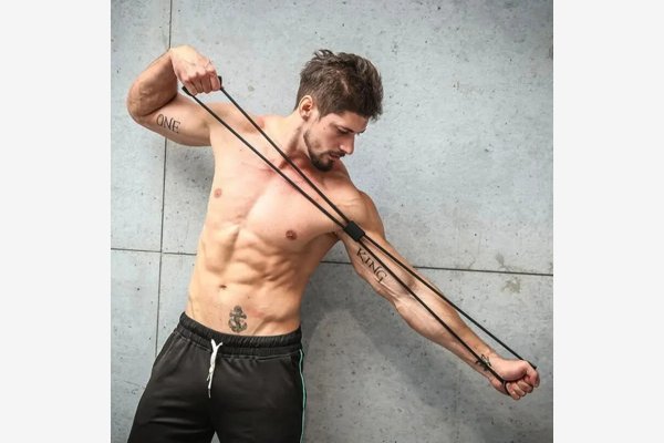 Homem fazendo exercício de braço aberto com o extensor de peito, em frente a uma parede de concreto