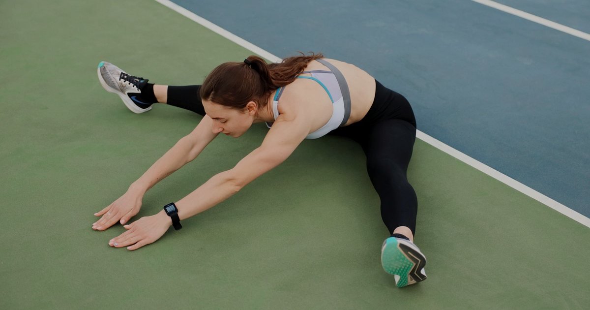 Mulher fazendo alongamento sentada em quadra, com as pernas abertas e o tronco inclinado à frente
