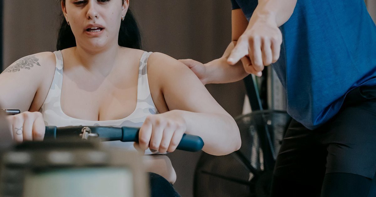 Mulher fazendo remada sentada em aparelho de academia, com um instrutor ao lado