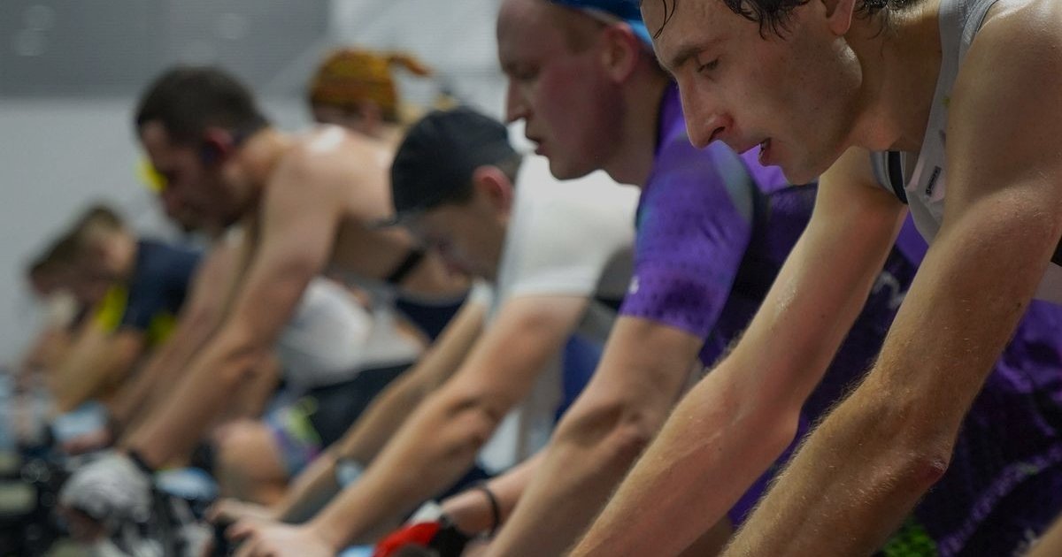 Grupo de pessoas pedalando lado a lado em uma aula de spinning, com expressões de esforço
