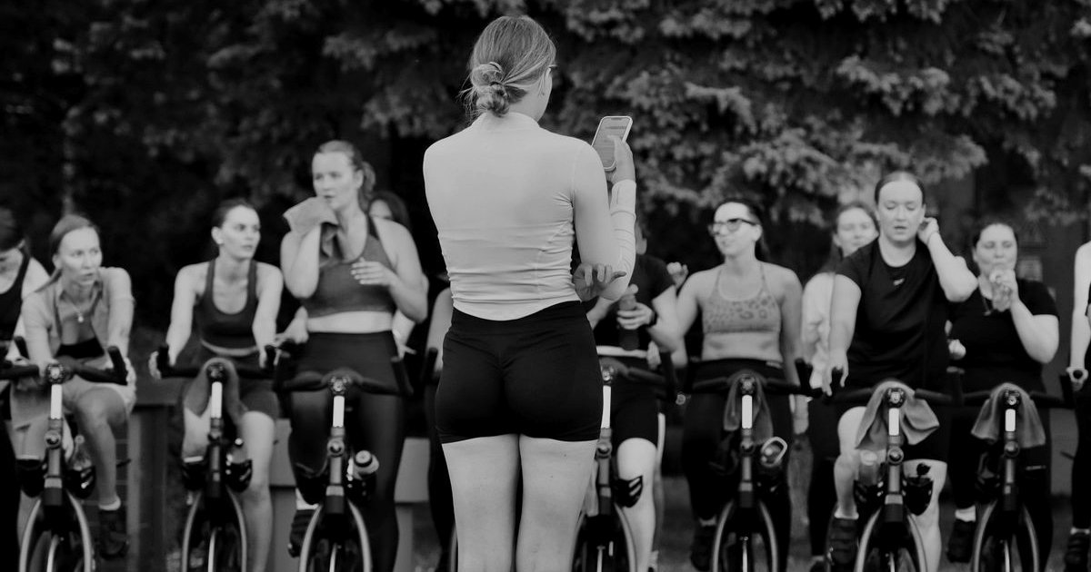 Aula de spinning ao ar livre em preto e branco, com instrutora de costas e alunas pedalando em fila