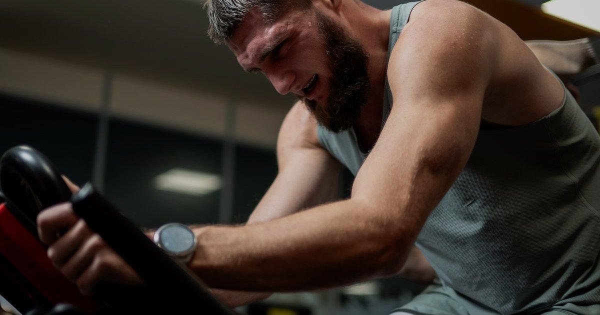 Homem de barba pedalando com esforço, mãos no guidão de uma bicicleta de academia