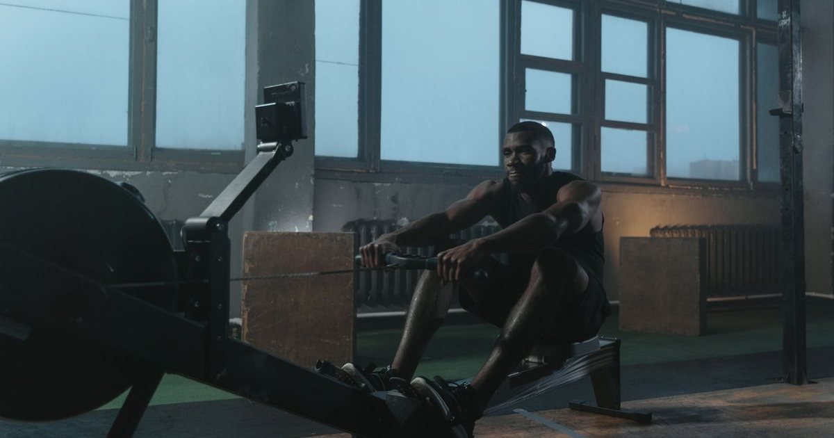 Atleta remando em uma máquina de remo indoor numa academia de janelas grandes